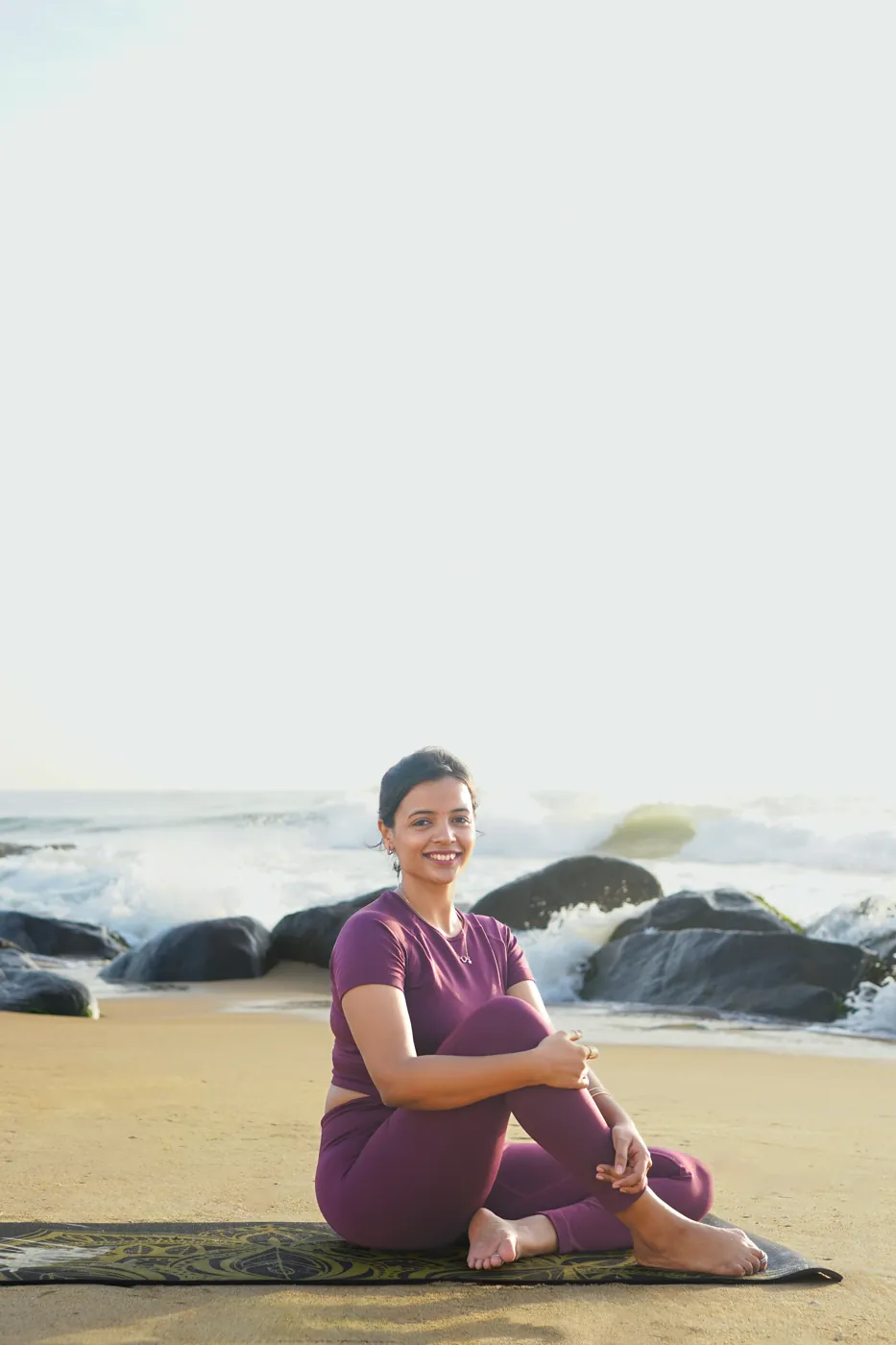 Ishwarya sitting on the sand and smiling, with the sea behind her